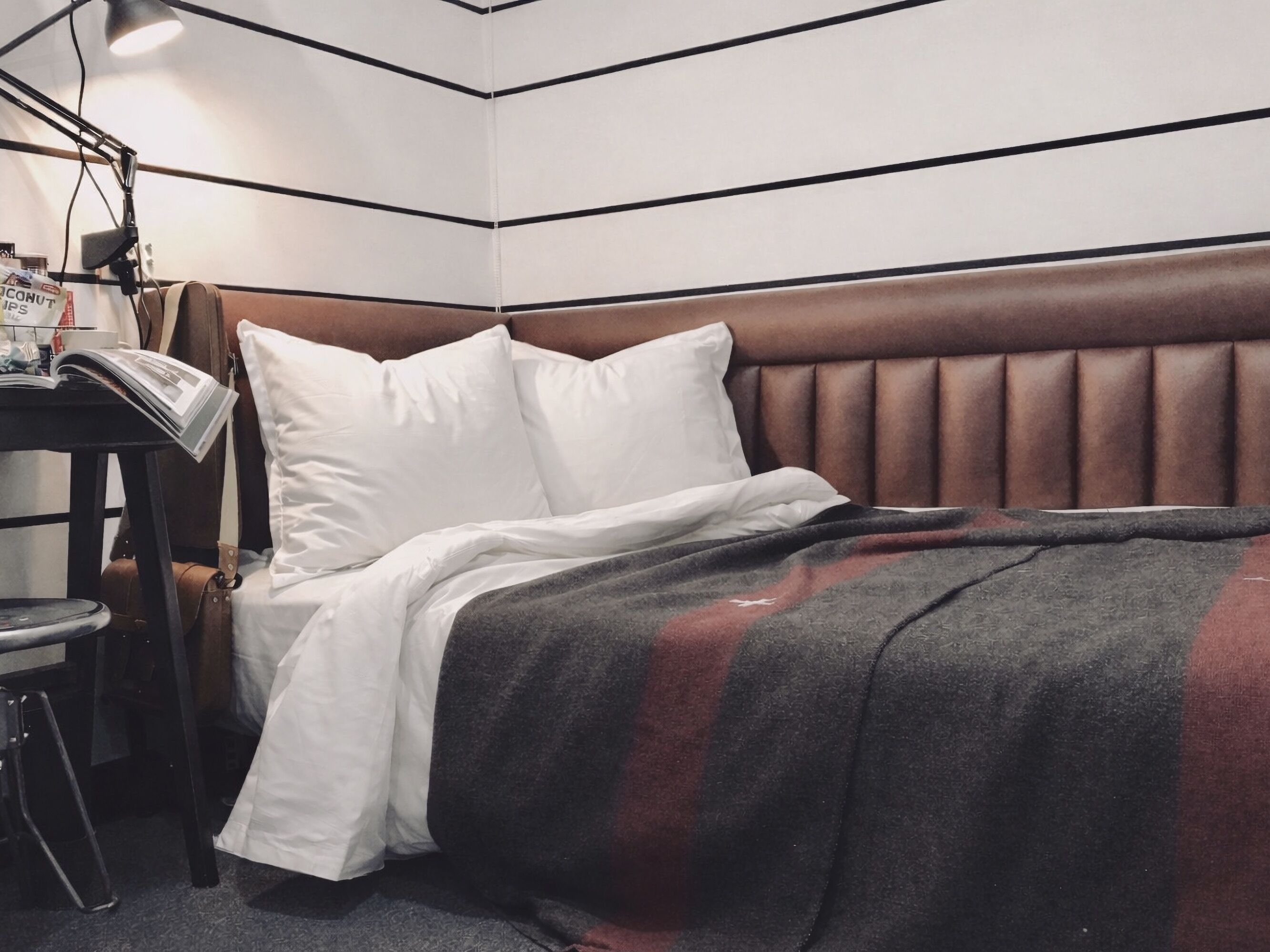 A neatly made bed with a brown headboard, white pillows, and a gray blanket. A small table with a lamp and a magazine is beside the bed.