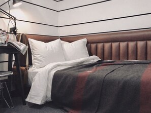 Een net opgemaakt bed met een bruin hoofdeinde, witte kussens en een grijze deken. Naast het bed staat een klein tafeltje met een lamp en een tijdschrift.