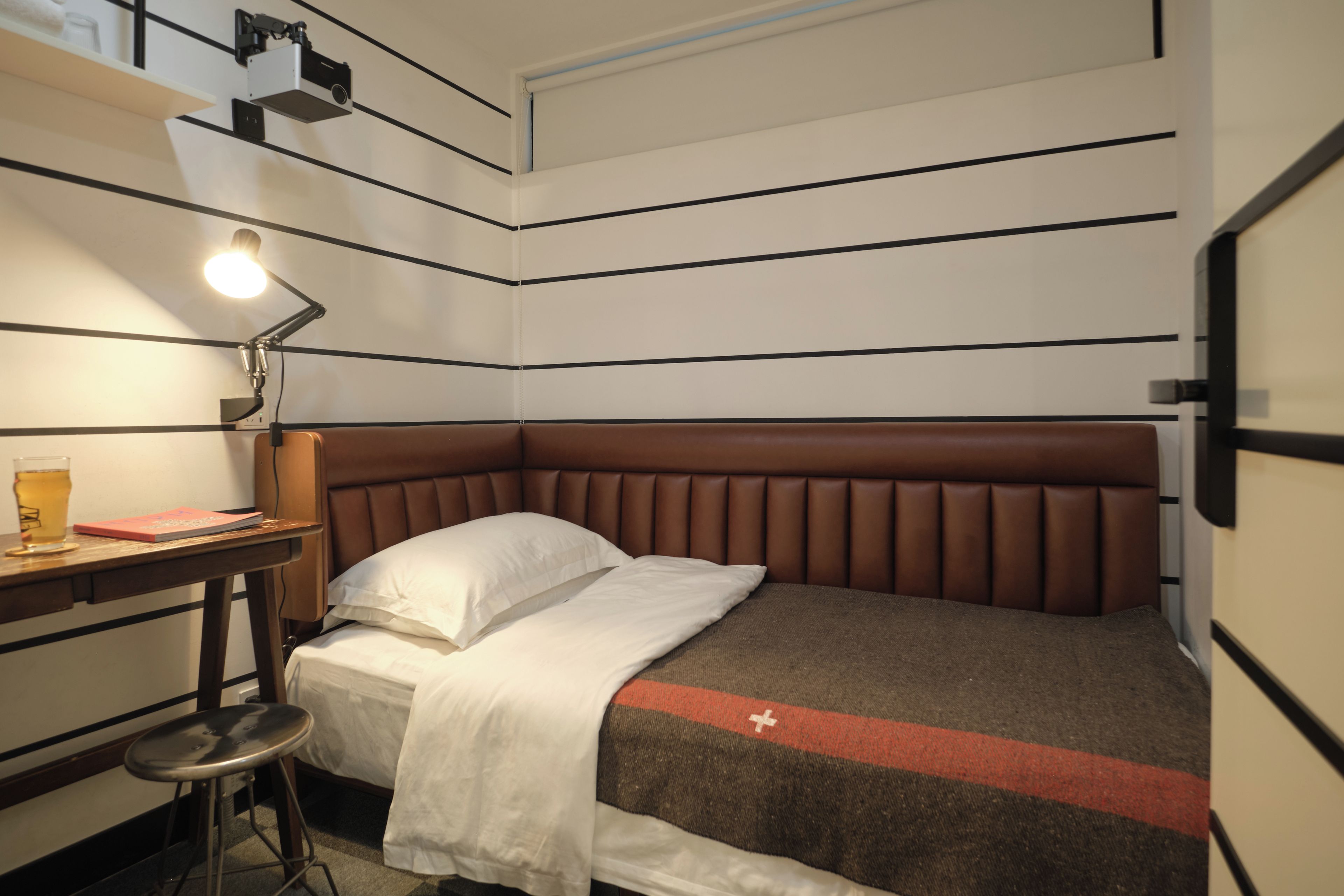 A compact hotel room with a single bed, a desk with a lamp, and a small stool.