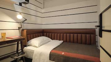 A compact hotel room with a single bed, a desk with a lamp, and a small stool.