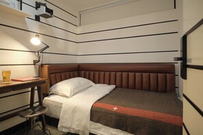 Une chambre d’hôtel de taille réduite, comprenant un lit simple, un bureau avec une lampe et un petit tabouret.