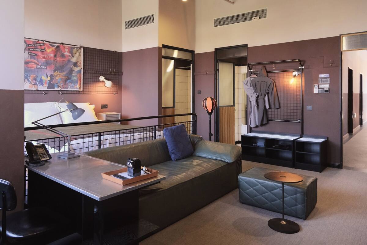 Een moderne hotelkamer met een bank, een klein tafeltje, een stoel, een bureau met telefoon, een lamp, een spiegel, een kapstok en een radiator.