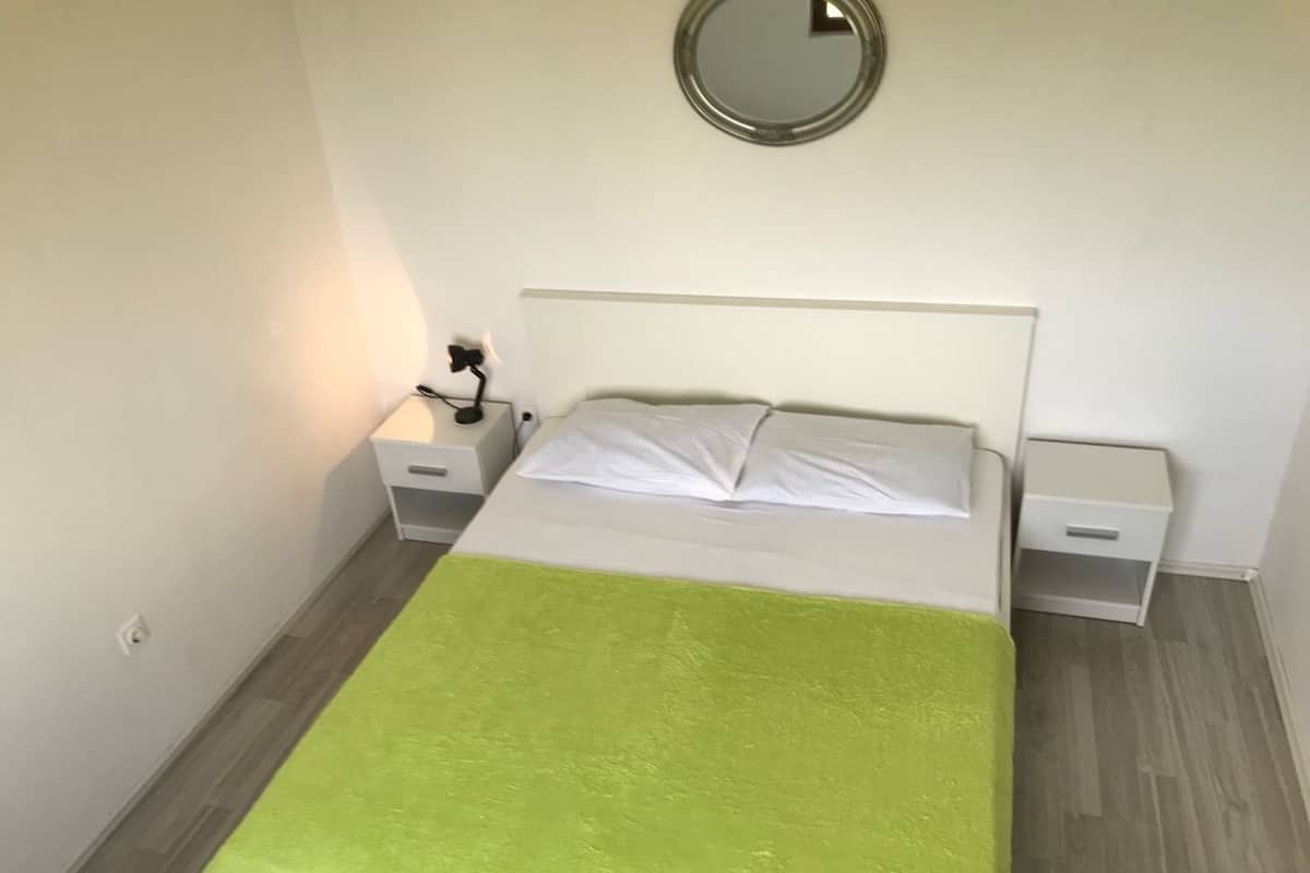 Ein ordentlich bezogenes Bett mit einer grünen Decke, zwei Nachttische mit Lampen und ein runder Spiegel an der Wand.