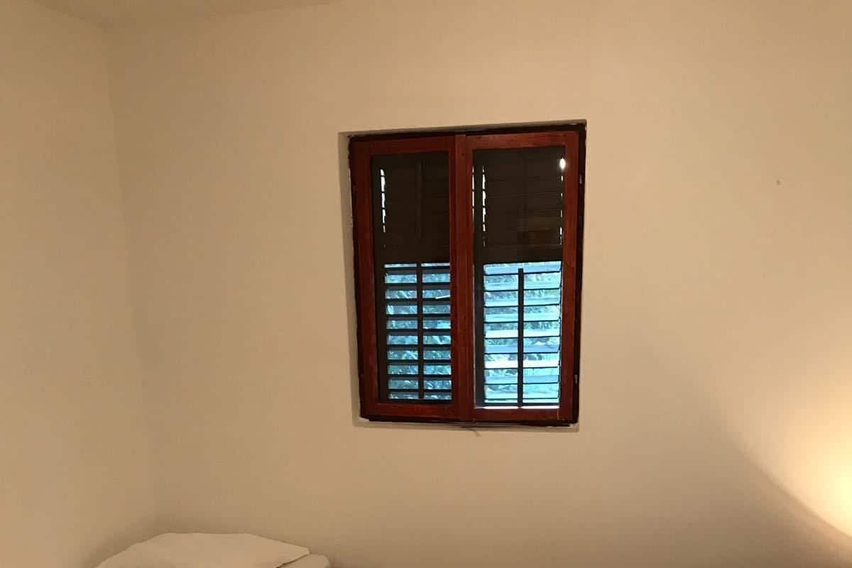 Ein kleiner, gemütlicher Raum mit zwei Einzelbetten, jeweils mit zusammengelegtem Handtuch und einem weißen Kopfkissen. Über den Betten befindet sich ein Fenster mit Jalousien, und der Boden ist mit Holz verlegt.