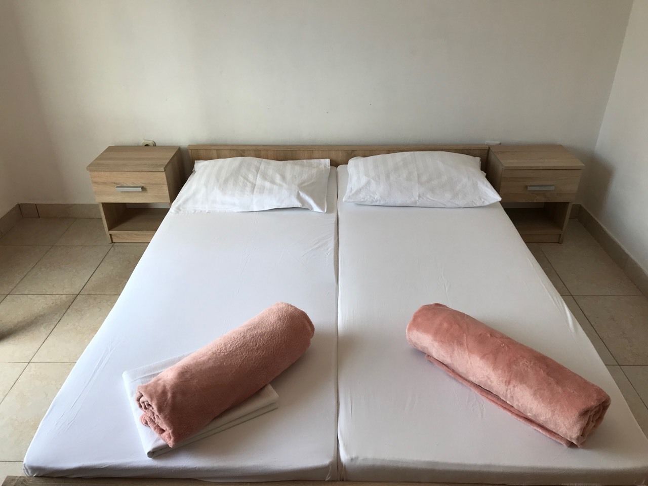 A double bed with white linens and two pink towels, wooden headboards, and bedside tables with drawers.
