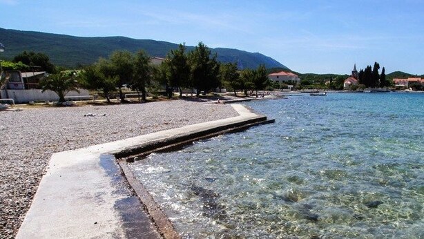 A pebble beach with clear water, a concrete walkway, and buildings in the background. - Sreser Dubrovnik Apartments Urlic (Janjina)