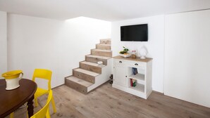 Una habitación pequeña con una escalera de madera, una mesa de comedor con sillas amarillas, un armario blanco con un televisor encima y una estantería.