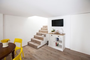 Una habitación pequeña con una escalera de madera, una mesa de comedor con sillas amarillas, un armario blanco con un televisor encima y una estantería.