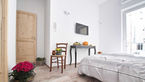 Un dormitorio con cama, suelo de madera, una silla, una mesita con frutas y una calabaza, y un televisor en la pared.