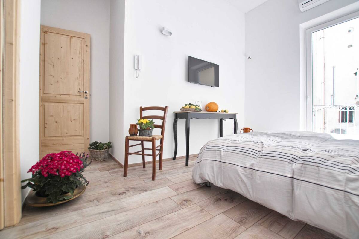 Ein Schlafzimmer mit Bett, Holzboden, einem Sessel, einem kleinen Tisch mit Früchten und einer Kürbis sowie einem Fernseher an der Wand.