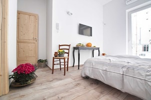 Un dormitorio con cama, suelo de madera, una silla, una mesita con frutas y una calabaza, y un televisor en la pared.