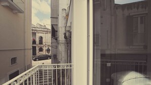 Una calle estrecha con un coche negro estacionado a la izquierda, un edificio blanco con un balcón a la derecha, y una cama con colchón blanco en primer plano.