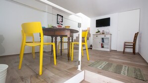 Un comedor con una mesa de madera y cuatro sillas amarillas, un armario blanco con un televisor encima, y una escalera que lleva a otro nivel.