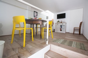 Un comedor con una mesa de madera y cuatro sillas amarillas, un armario blanco con un televisor encima, y una escalera que lleva a otro nivel.