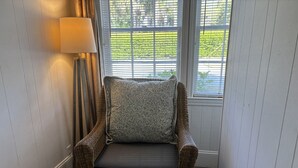 A wicker armchair with a cushion and a lamp in a corner of a room with white paneled walls and a window with blinds.