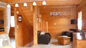 Interior d'una cabana de fusta amb una escala, una zona d'estar amb un sofà i cadires, una taula de menjador i una cuina.