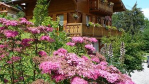 Una xalet de fusta amb balcons i caixes de flors, envoltada de vegetació abundant i flors roses.