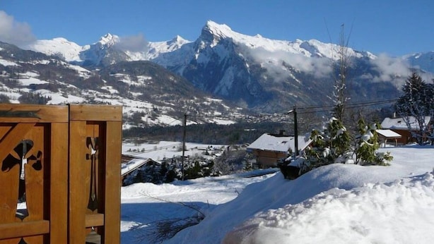 Un pont de fusta amb vistes a muntanyes cobertes de neu i una casa petita al fons.