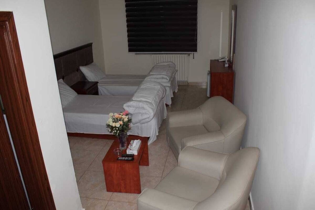 A hotel room with two beds, a sofa, a wooden coffee table with a vase of flowers, and a bedside table with a lamp.