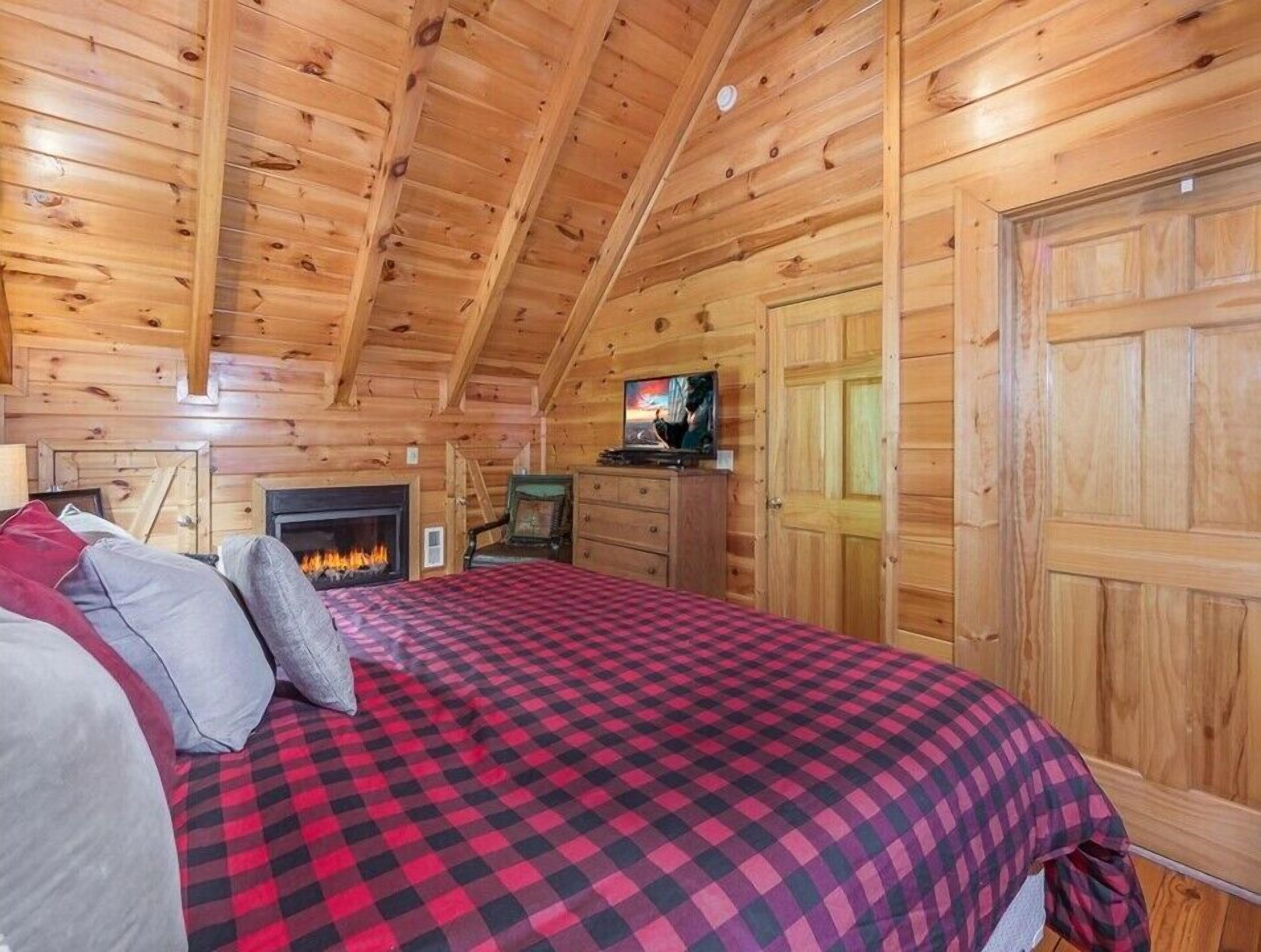 Une chambre confortable dans un chalet en rondins, avec une couverture à carreaux sur le lit, une cheminée, un téléviseur et des murs en bois.