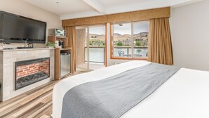 A hotel room with a large bed, a fireplace, a television, a kitchen area, and a balcony with a view of the mountains. - GrandView 723- Waterfront King View River View (Chelan)