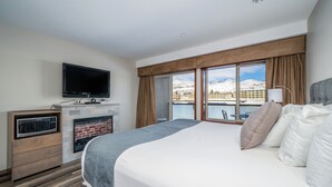 A hotel room with a large bed, a flat-screen TV, a fireplace, and a view of a snowy mountain and water.