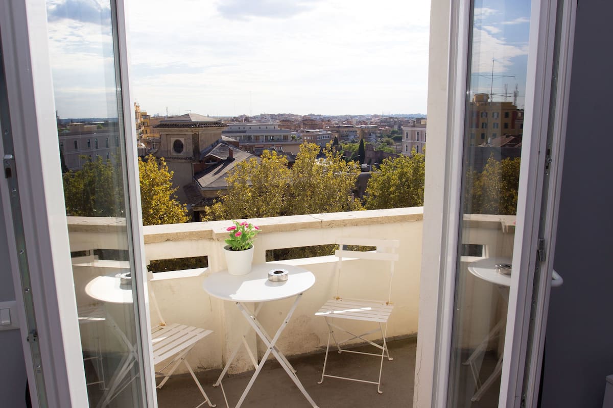 En balkon med et hvidt bord og en stol, en potteplante og udsigt over byen.