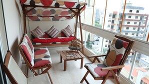 A balcony with a wooden swing bed, two chairs, a small table, and a view of the cityscape. - Vino Apartment (Zalqa)