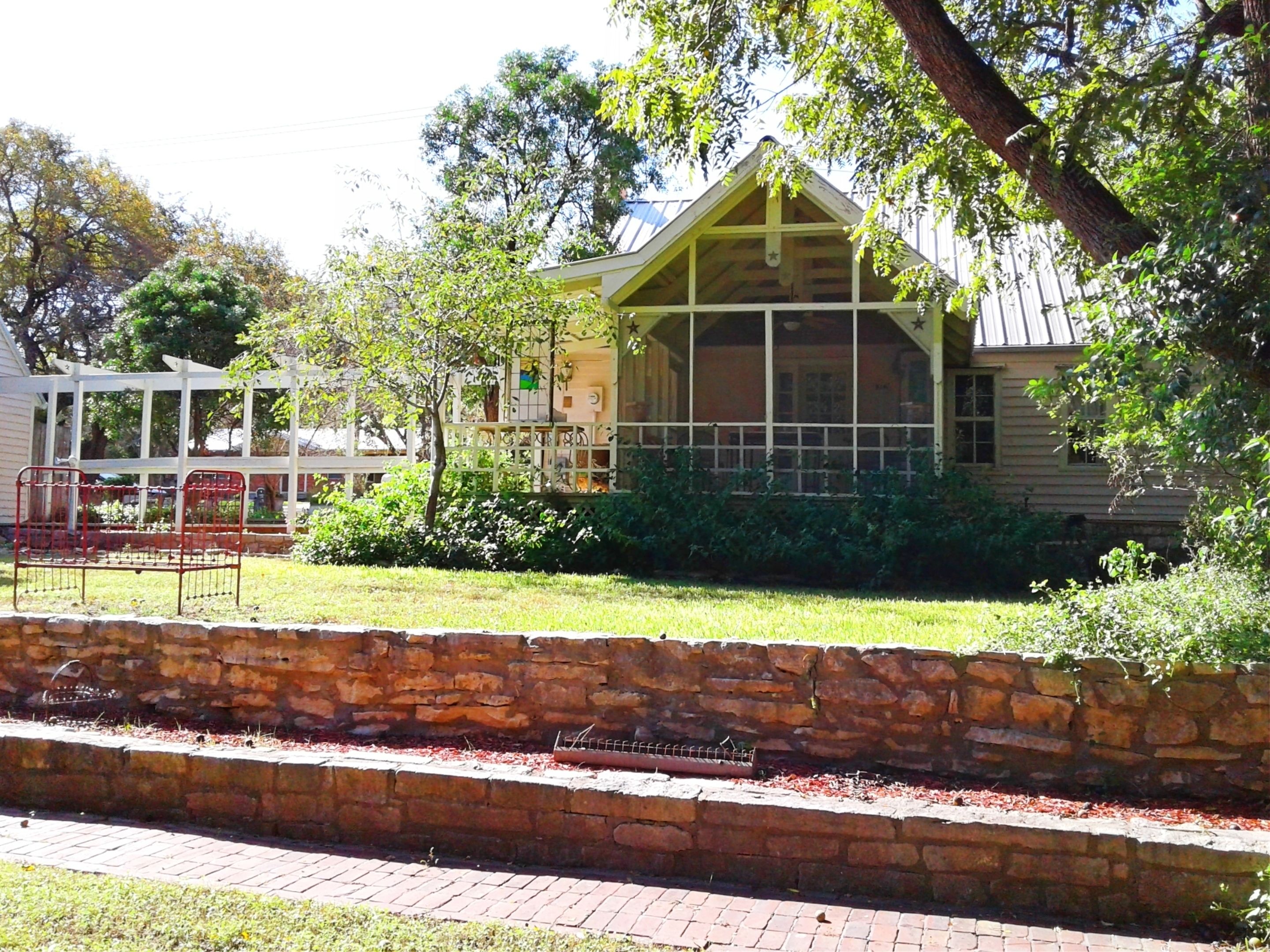 A rustic wooden building with a porch, surrounded by greenery and a stone wall.
