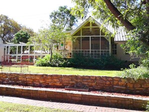 A rustic wooden building with a porch, surrounded by greenery and a stone wall.