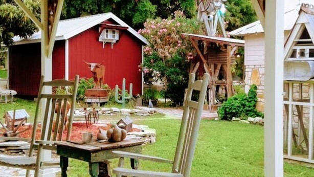 A rustic outdoor dining area with a wooden table and chairs, surrounded by a garden with a red barn and various plants. - The Baines House Inn (Salado)
