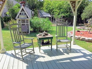 A wooden deck with two rocking chairs and a small table, surrounded by a garden and a gazebo.