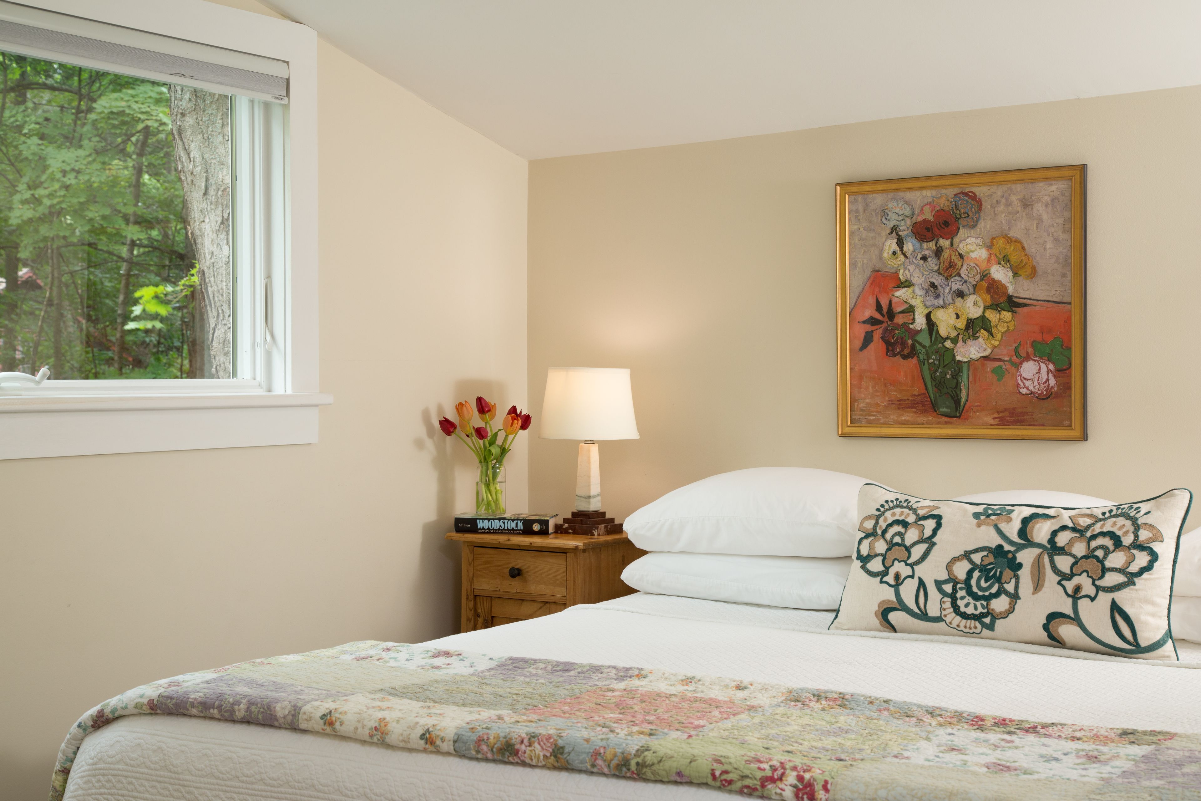 A neatly made bed with a floral quilt, a bedside table with a lamp, a vase of flowers, and a painting of flowers on the wall.