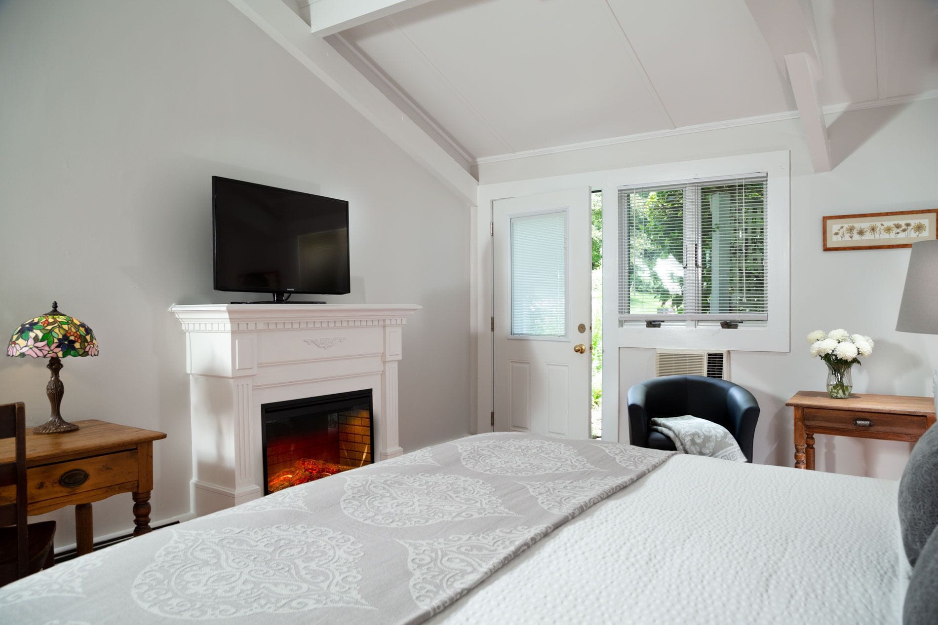 A bedroom with a fireplace, a television on a mantel, a bed with a patterned bedspread, a chair, and a nightstand with a lamp.