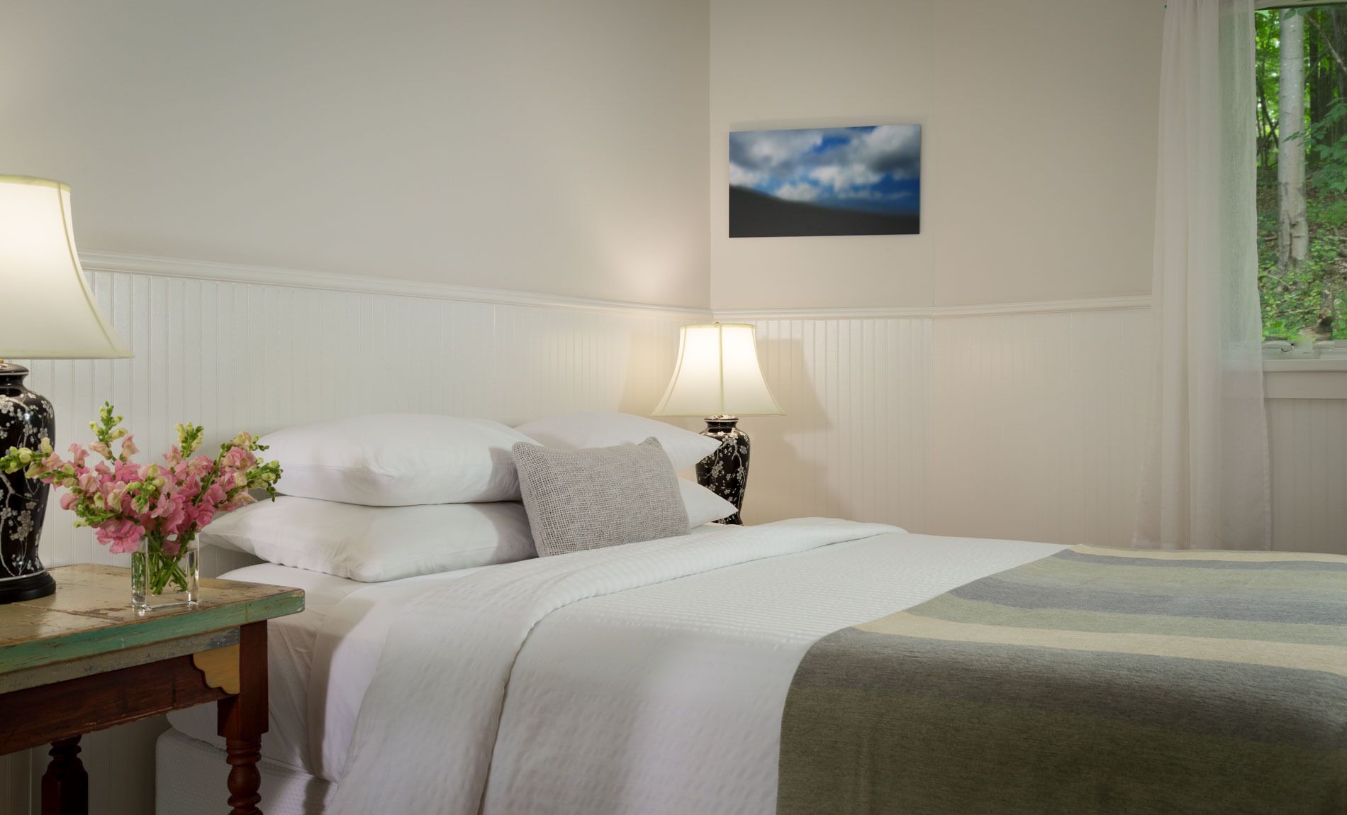 A neatly made bed with white linens, a vase of pink flowers, and two bedside lamps on a wooden nightstand.