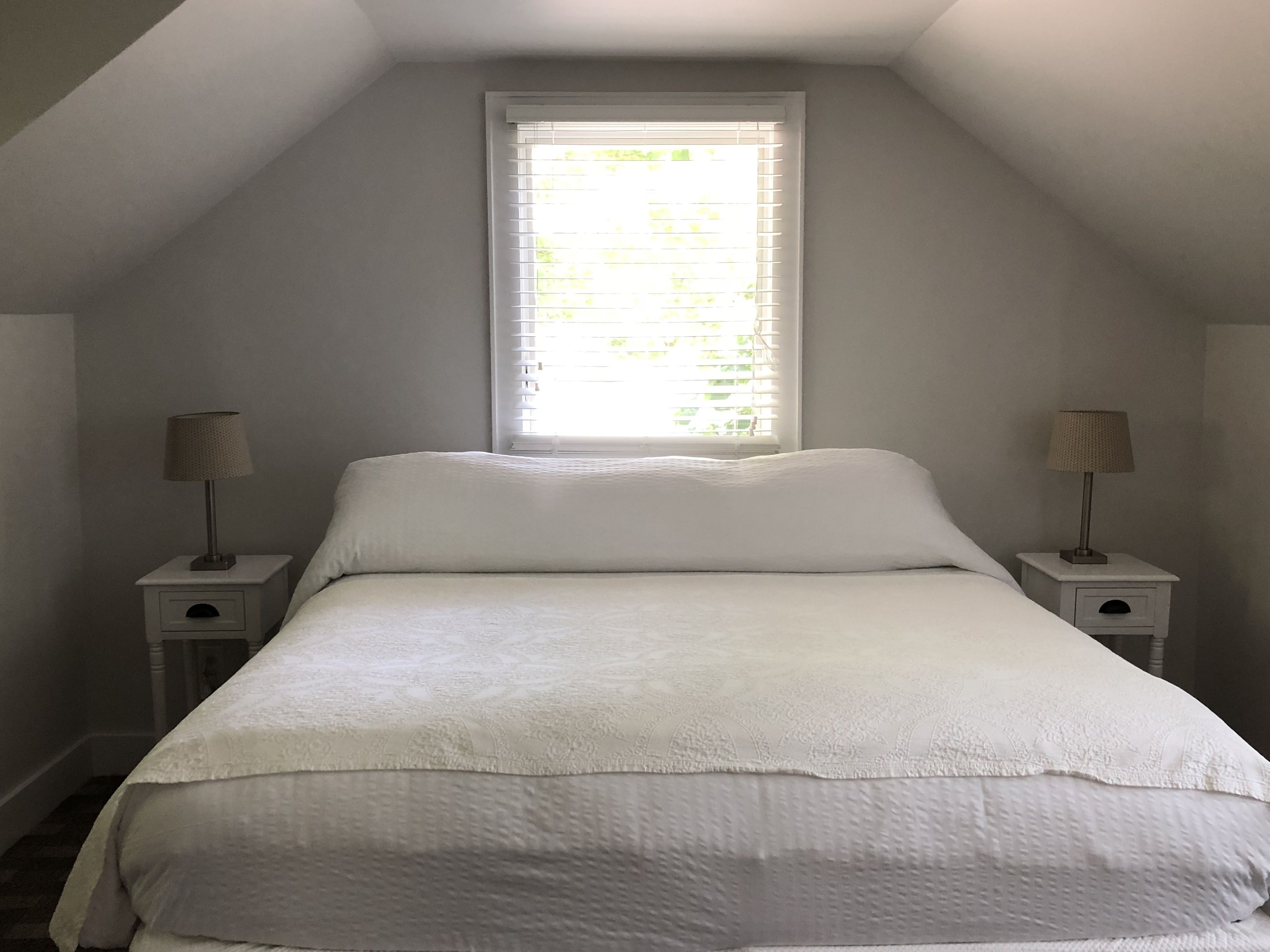 A bedroom with a sloped ceiling, a large window with blinds, a bed with white bedding, and two bedside tables with lamps.