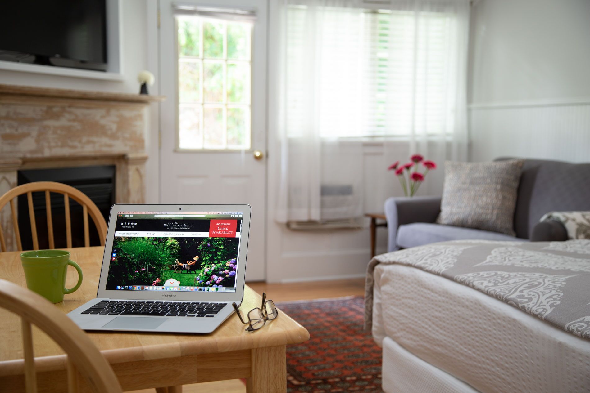 A laptop on a wooden table with a green mug, a bed with patterned bedding, a grey sofa, and a window with blinds.