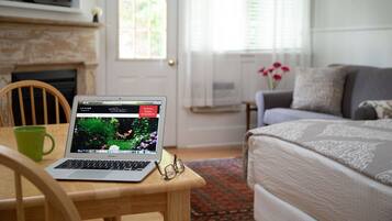 A laptop on a wooden table with a green mug, a bed with patterned bedding, a grey sofa, and a window with blinds.