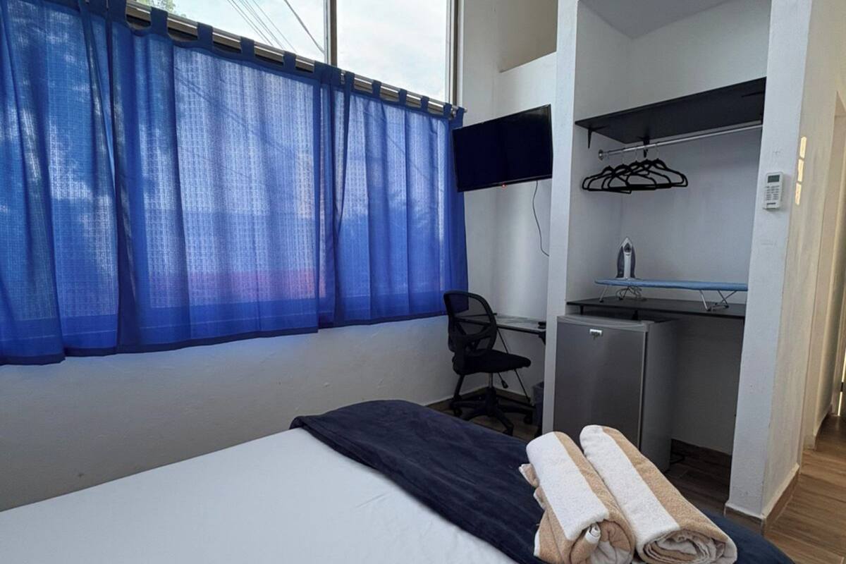 Una habitación de hotel con cama, escritorio, silla, televisión y una kitchenette pequeña.
