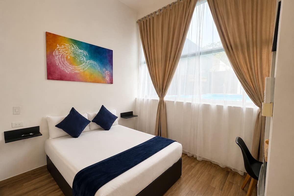 Habitación de hotel con cama, mesita de noche, escritorio, silla, un cuadro y ventana con cortinas.