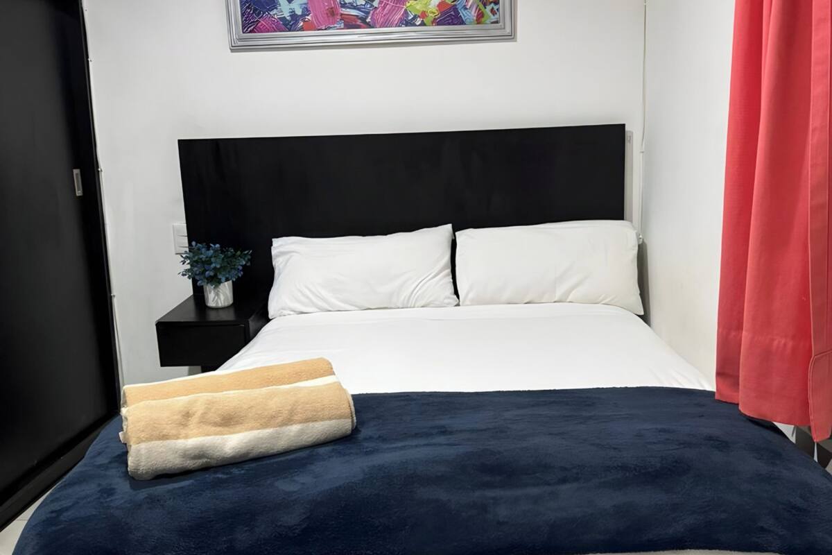 Una cama bien hecha con ropa de cama blanca y azul marino, una manta color beige y una mesita de noche con una pequeña planta.