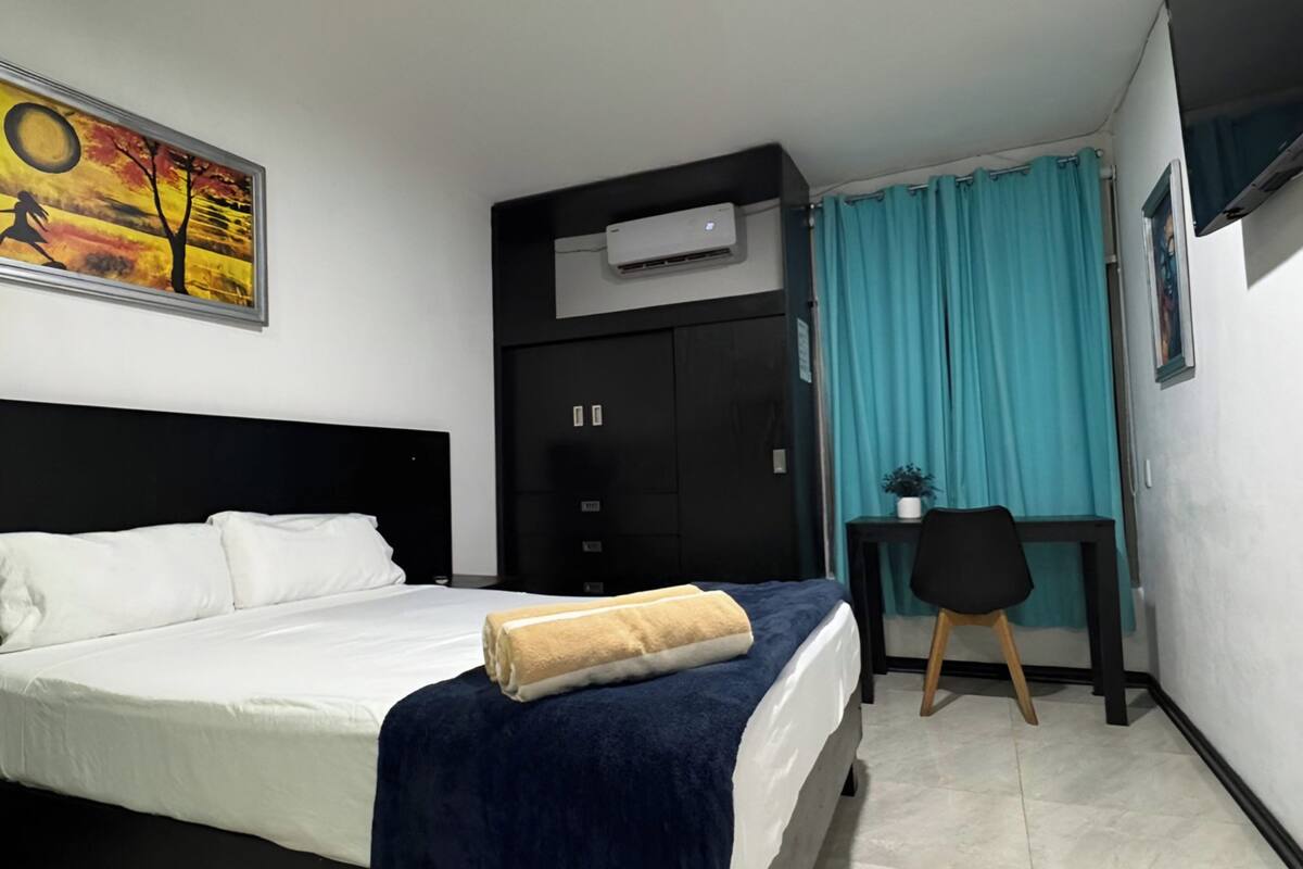Una habitación de hotel con una cama, un escritorio con una silla, un equipo de aire acondicionado y un cuadro en la pared.
