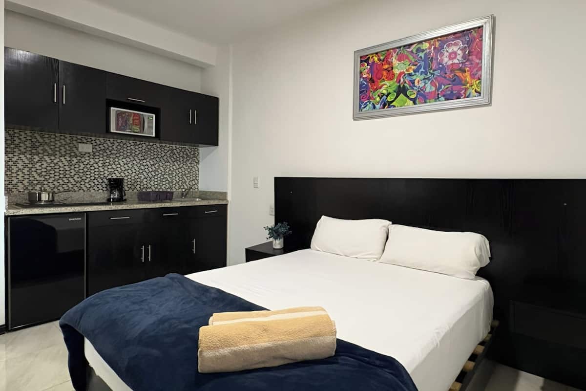 Un dormitorio moderno con una cama grande, una mesita de noche pequeña, una mesita auxiliar y una zona de cocina con armarios oscuros y un salpicadero de azulejos.