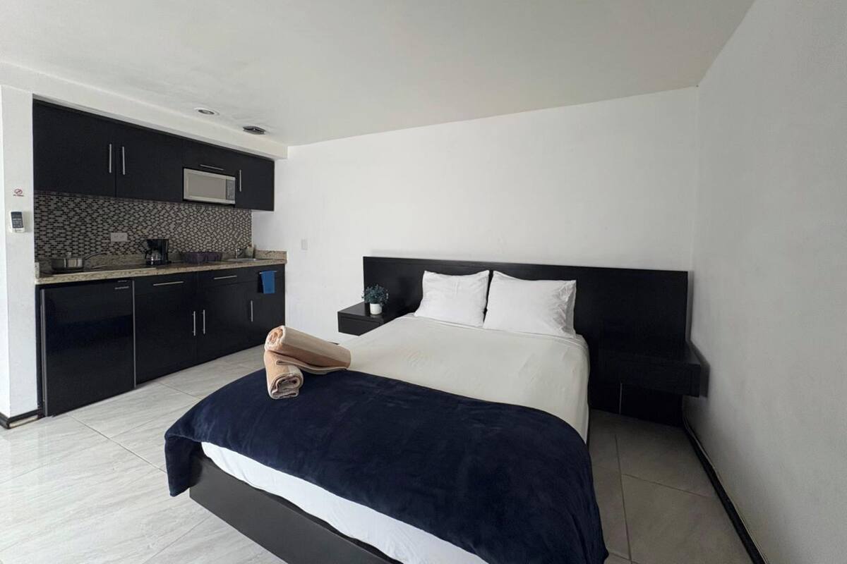Un dormitorio moderno con una cama grande, una mesita de noche pequeña y una cocina integrada con armarios oscuros y un salpicadero de azulejos.