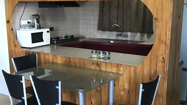 Cocina compacta con pared de madera, microondas blanco, un lavamanos y una mesa de vidrio con cuatro sillas.