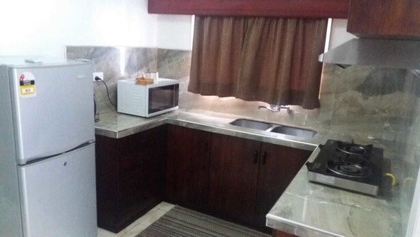 A compact kitchen with dark wooden cabinets, a white refrigerator, a microwave, a sink, and a gas stove. - Two Bedroom House HHK-17-2 (Lautoka)