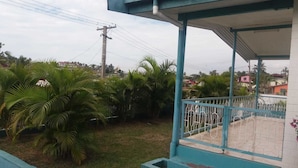 A covered outdoor area with a metal railing, surrounded by greenery and a view of a residential area in the background. - Two Bedroom House HHK-17-2 (Lautoka)
