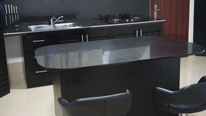 A modern kitchen with a central island, black cabinets, stainless steel appliances, and two black bar stools. - One Bedroom House HHK-19-1 (Lautoka)