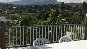 A balcony with a white table and chairs, overlooking a residential area with houses and mountains in the background. - One Bedroom House HHK-19-1 (Lautoka)