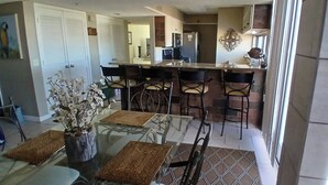 Un comedor con mesa y sillas de vidrio, un florero con flores y una cocina al fondo.